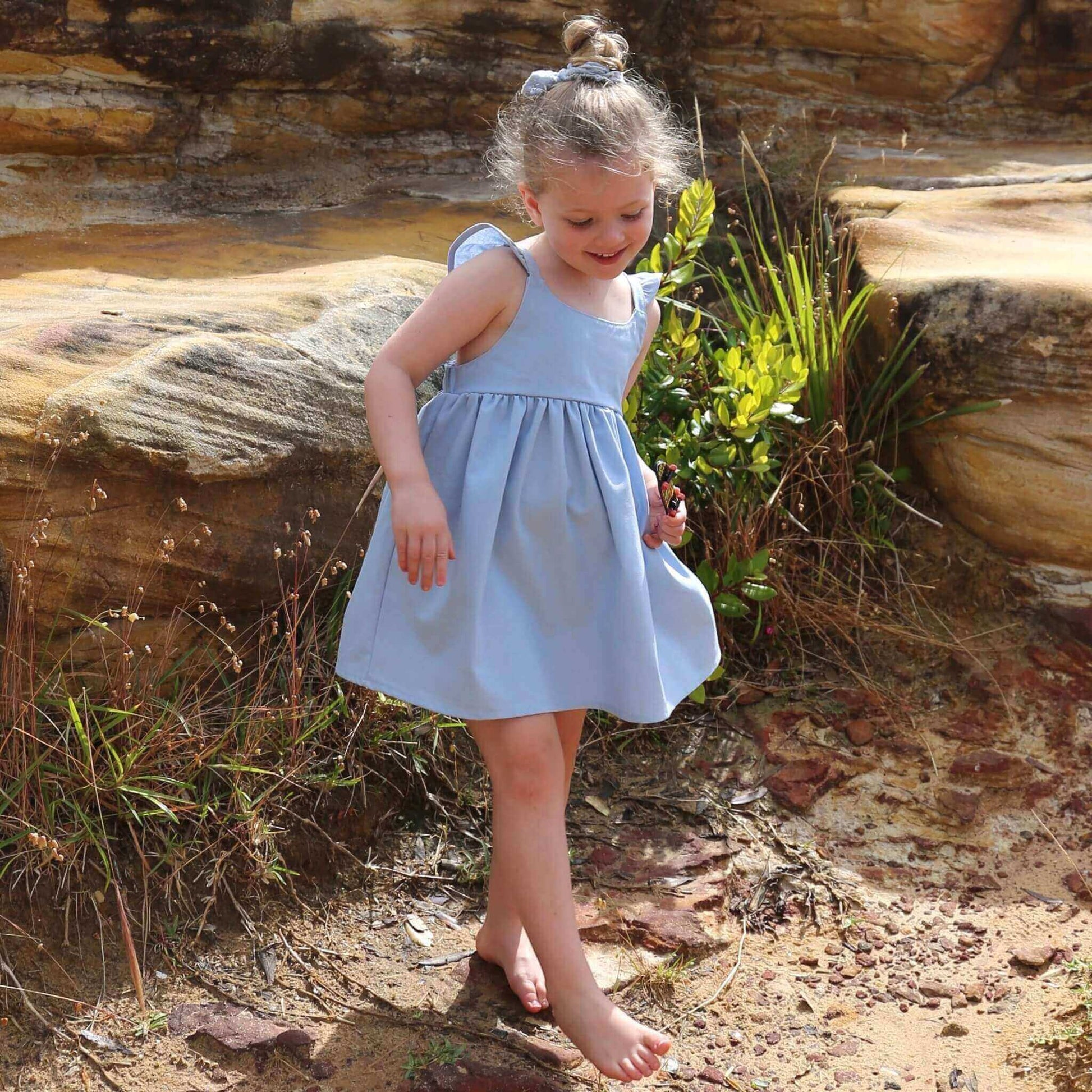 Girl wearing Sky Blue Dress featuring a gathered skirt and frill on back crossed straps. Ethically made in Australia with a Linen Cotton blend fabric