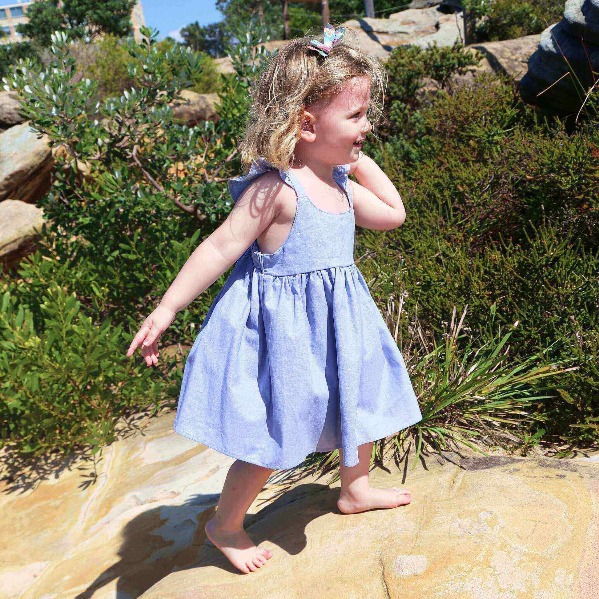 Girl wearing Blue Dress featuring a gathered skirt and frill on back crossed straps. Ethically made in Australia with 100% Cotton Chambray fabric