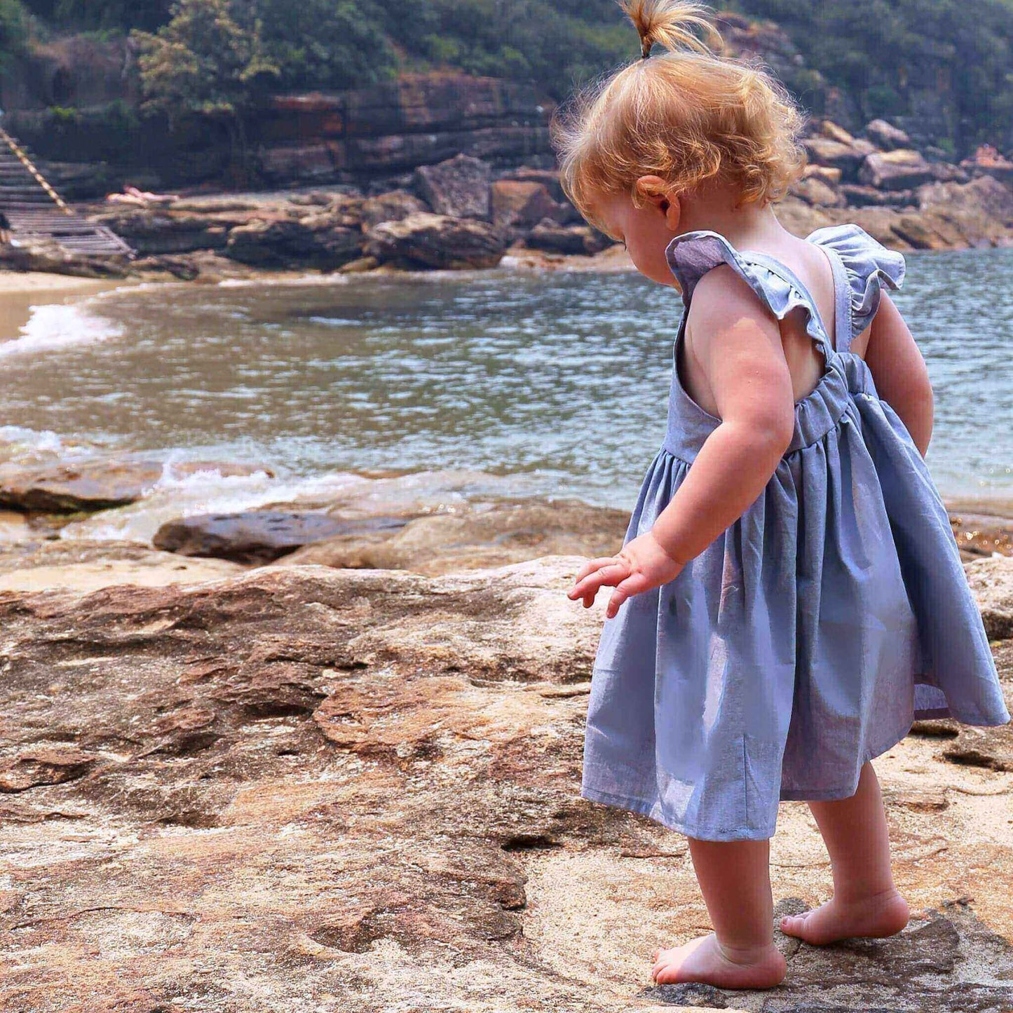 Toddler Girl wearing Blue Dress featuring a gathered skirt and frill on back crossed straps. Ethically made in Australia with 100% Cotton Chambray fabric