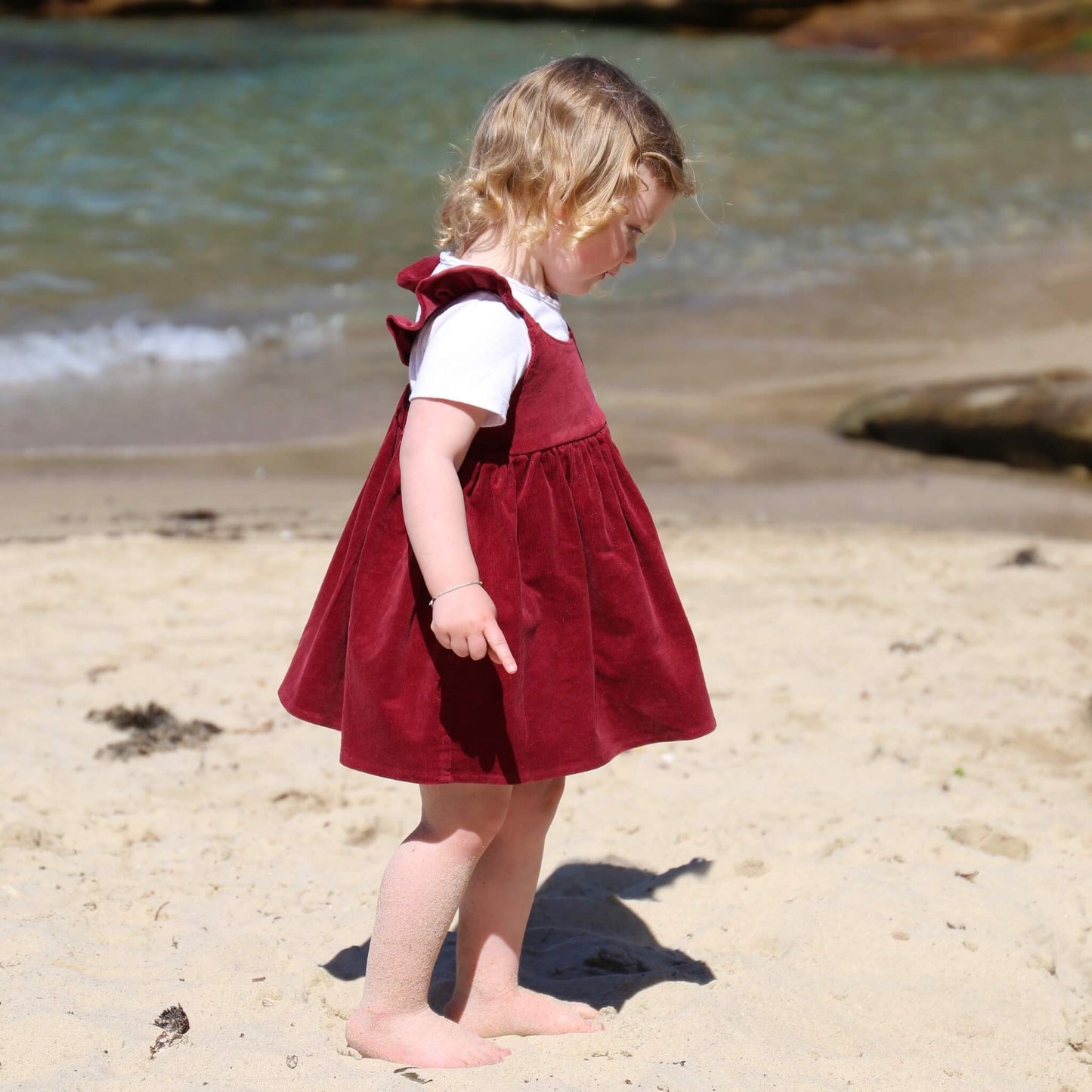 Toddler girl wearing Ruby coloured Dress featuring a gathered skirt and frill on back crossed straps. Ethically made in Australia with a Cotton Elastane corduroy fabric