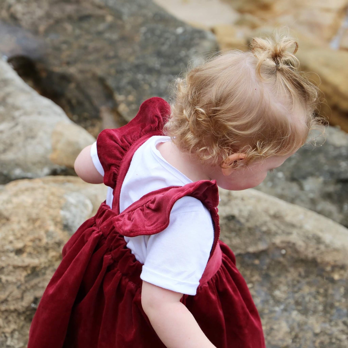 Toddler girl wearing Ruby coloured Dress featuring a gathered skirt and frill on back crossed straps. Ethically made in Australia with a Cotton Elastane corduroy fabric