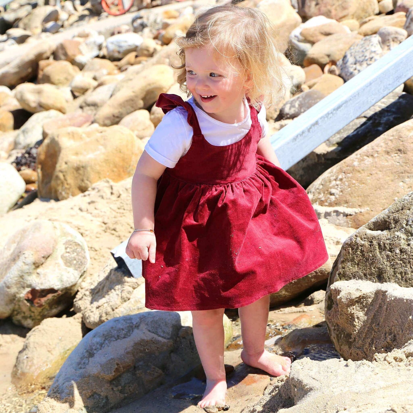 Toddler girl wearing Ruby coloured Dress featuring a gathered skirt and frill on back crossed straps. Ethically made in Australia with a Cotton Elastane corduroy fabric