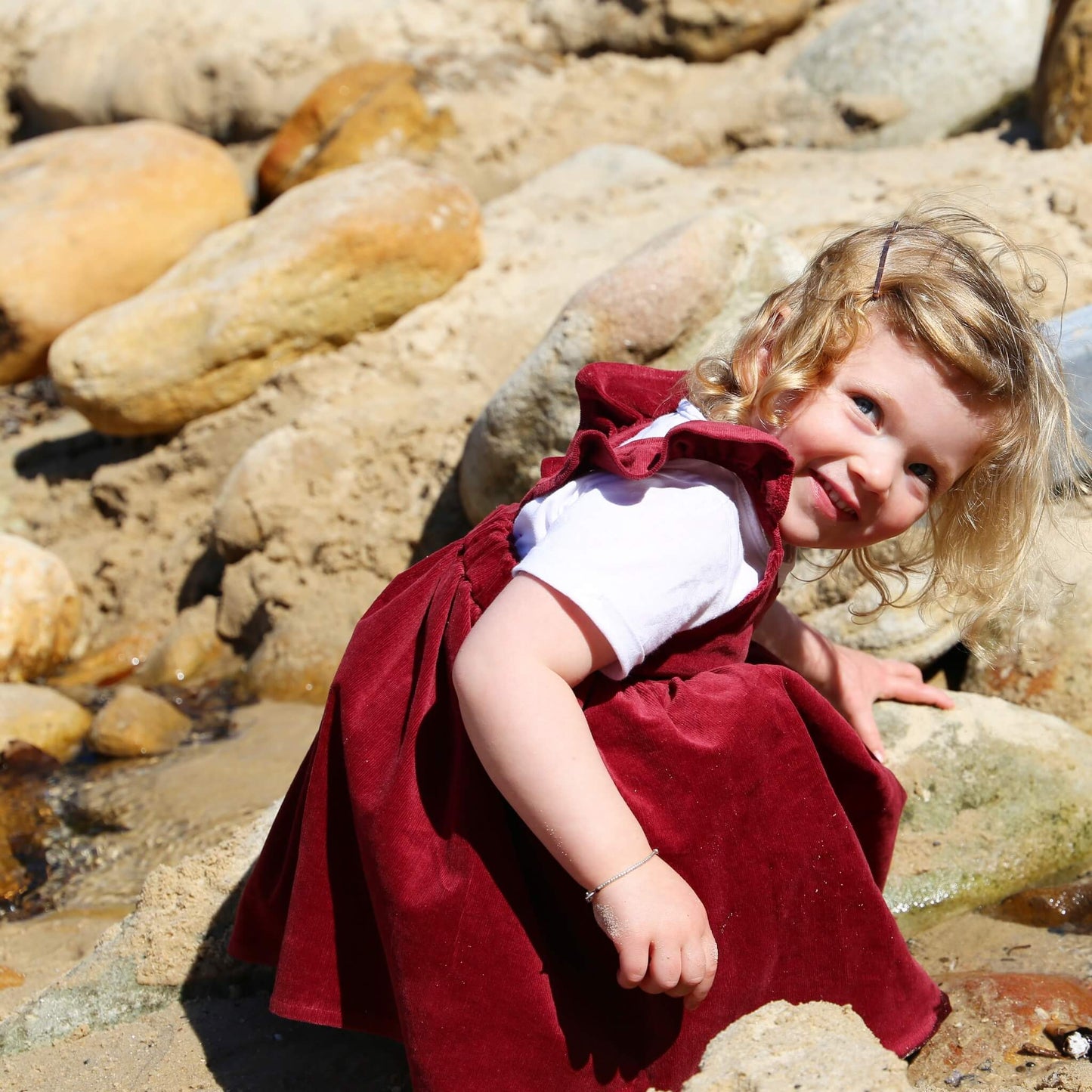 Toddler girl wearing Ruby coloured Dress featuring a gathered skirt and frill on back crossed straps. Ethically made in Australia with a Cotton Elastane corduroy fabric