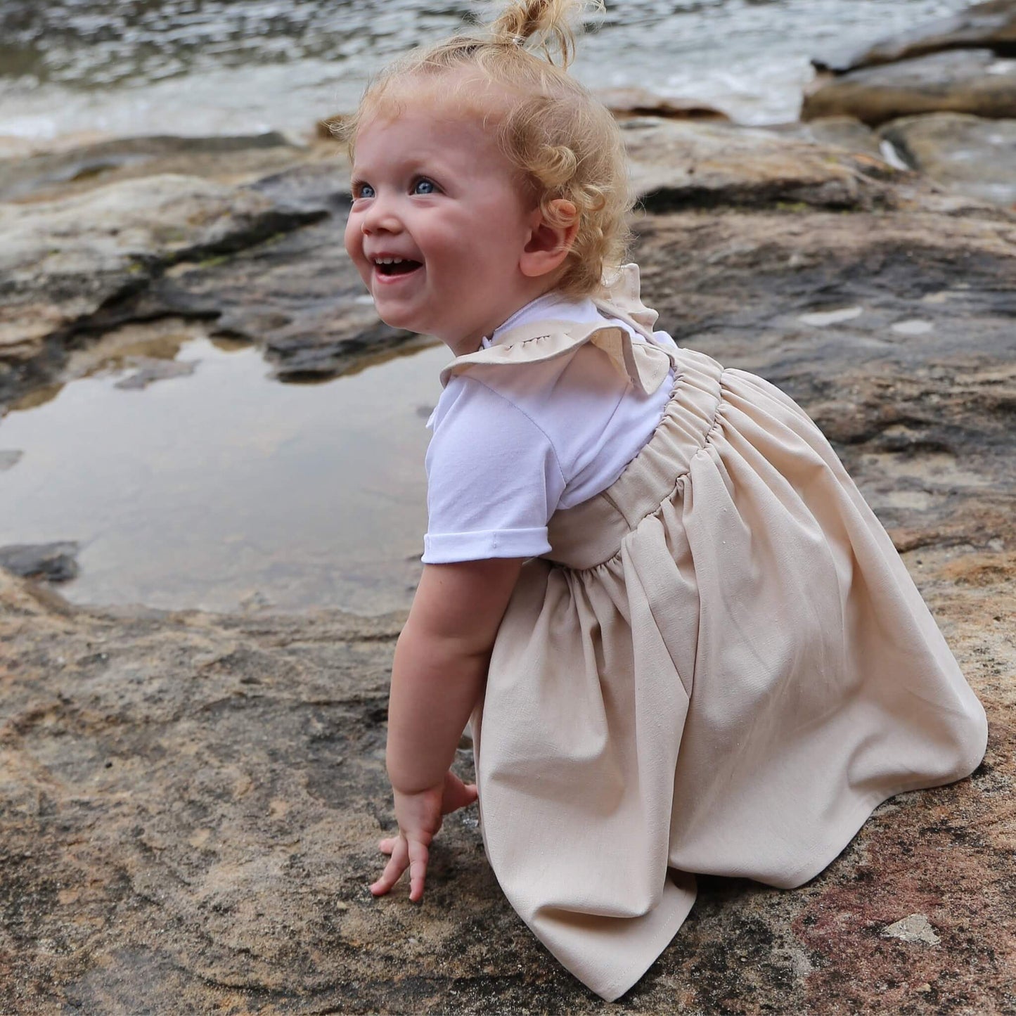 Toddler Girl wearing Linen colour Dress featuring a gathered skirt and frill on back crossed straps. Ethically made in Australia with a Linen Cotton blend fabric